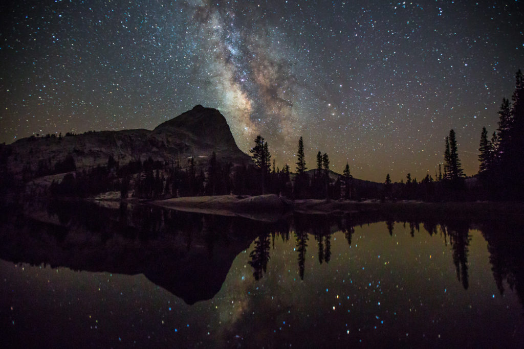 Milky Way stars reflected in an alpine lake beneath dark mountain silhouettes