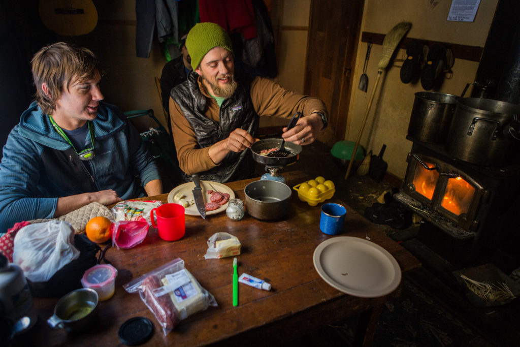 Two people sharing food beside a wood stove in a cabin