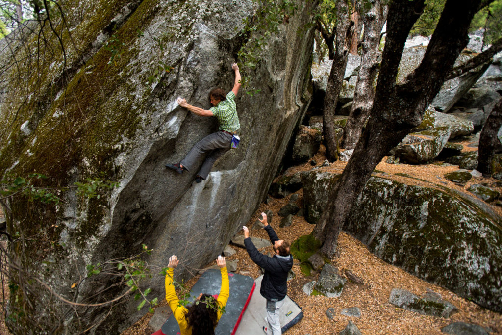 Boulderer in a green top climbing above two spotters in the forest