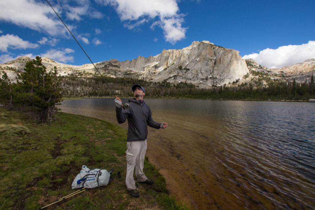 Fly fisher casting from a grassy alpine lakeshore beneath granite peaks
