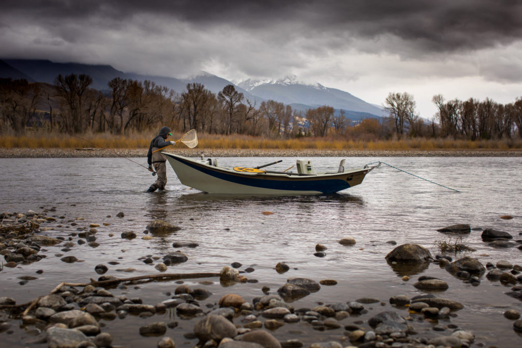 Angler beside a drift boat on a shallow river under storm clouds
