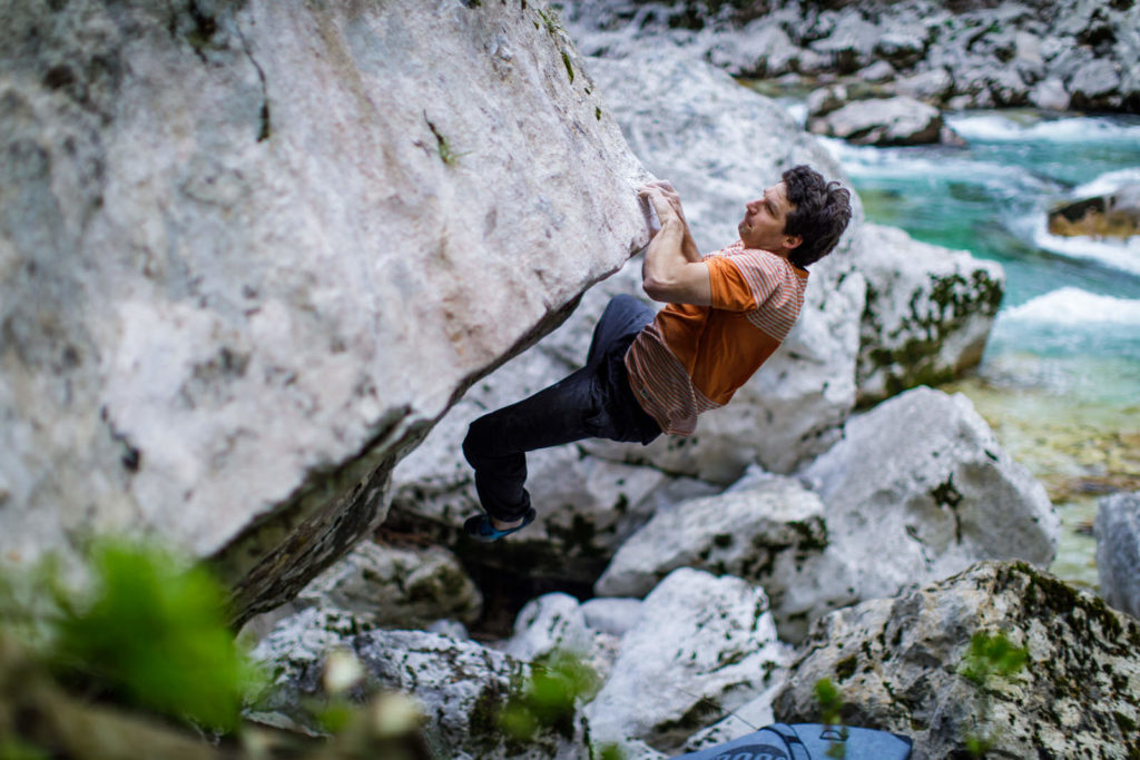 Climber in an orange shirt gripping an overhanging riverside boulder