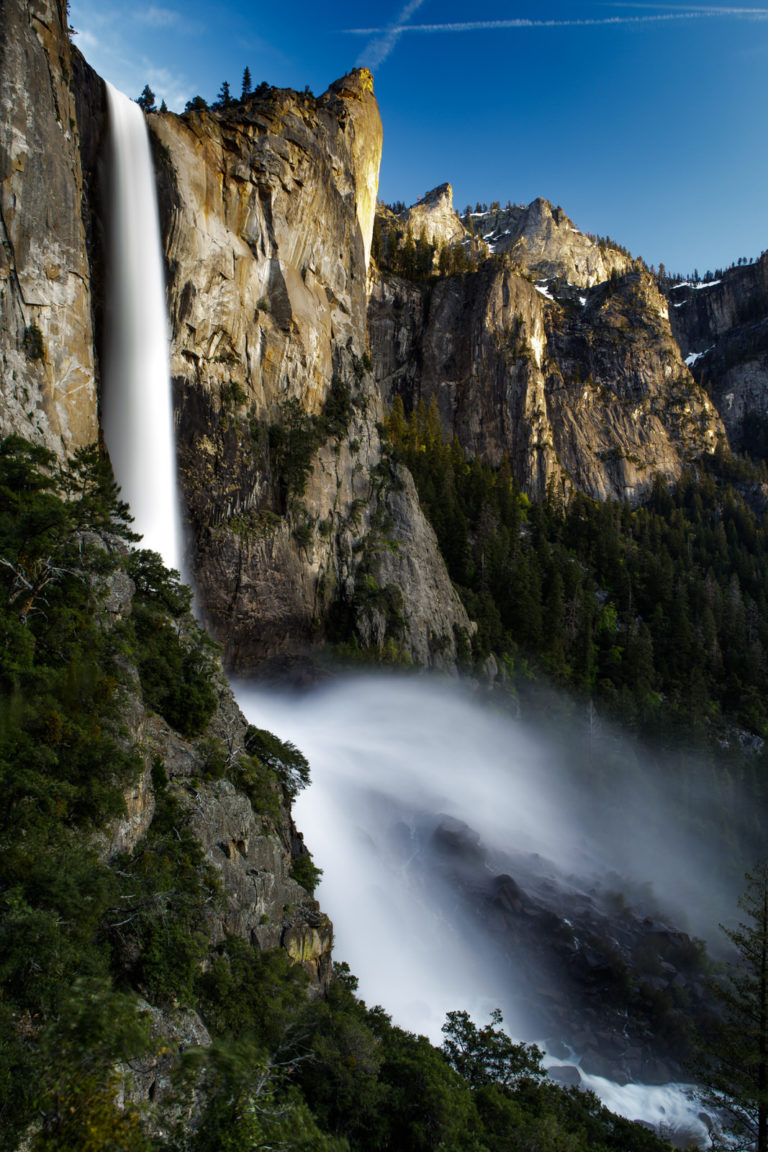Waterfall plunging past a sunlit granite cliff into a forested valley