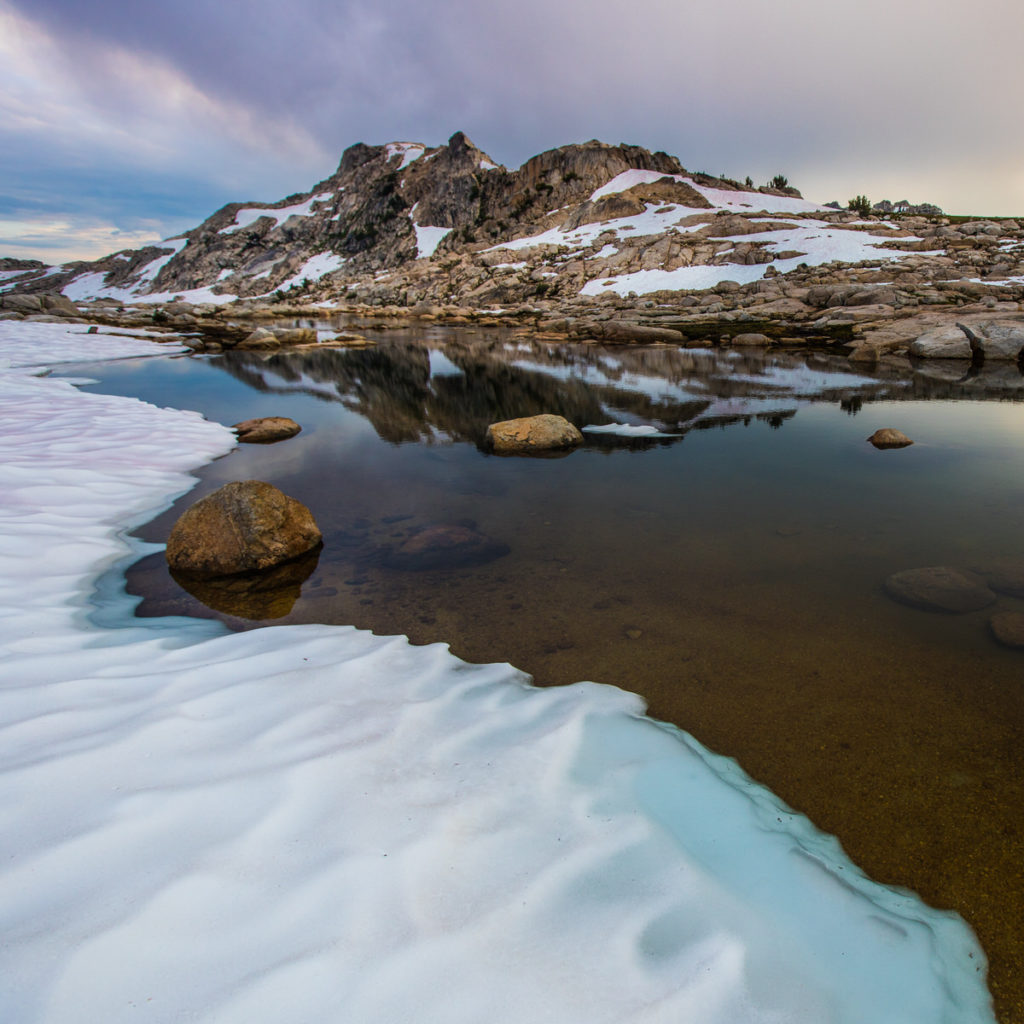 Snowbanks and granite boulders reflected in an alpine pool