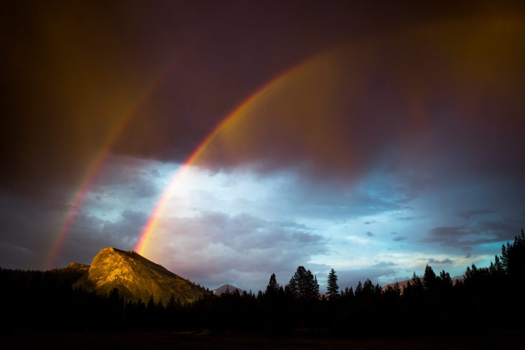 Double rainbow beneath dark storm clouds above a forested ridge
