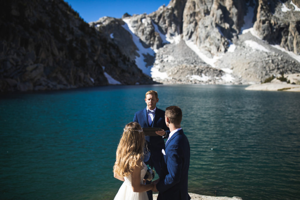 Couple exchanging vows with an officiant beside an alpine lake