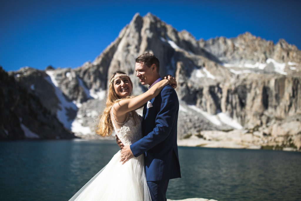 Newlyweds smiling and embracing beside an alpine lake