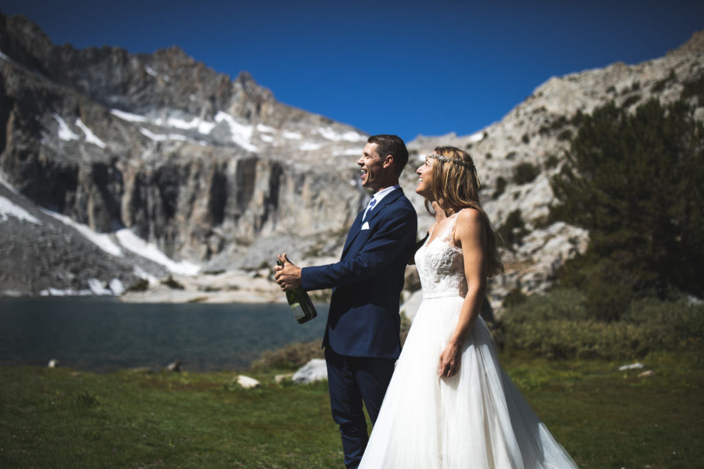 Groom opening a bottle beside the bride at a mountain lake