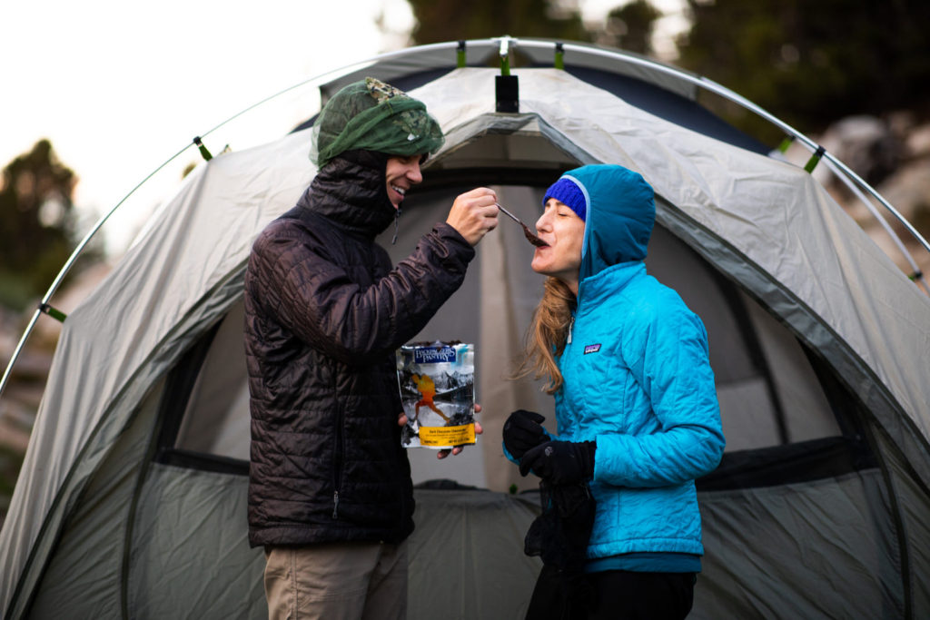 Couple sharing a snack outside their tent in warm hiking jackets