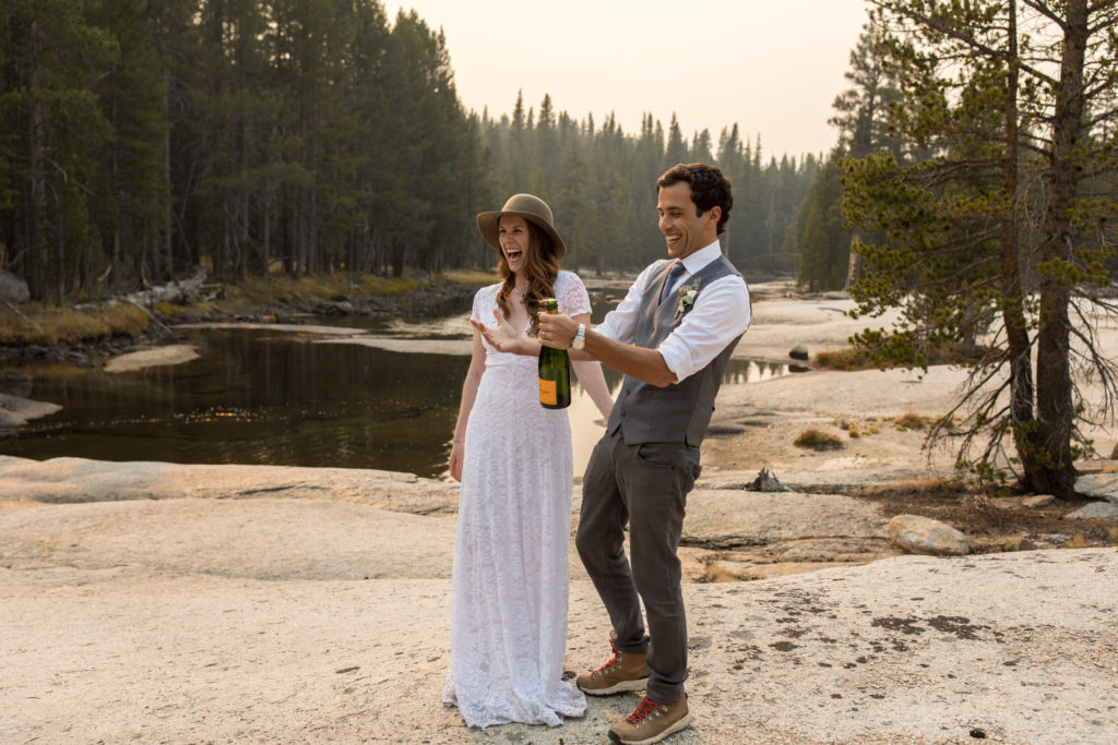 Groom opening a bottle beside the bride near a forest lake