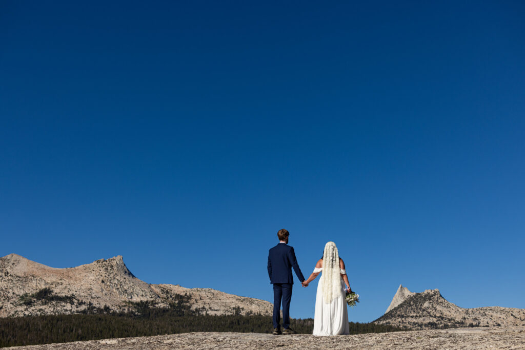 Bride and groom holding hands on a mountain ridge beneath a clear blue sky