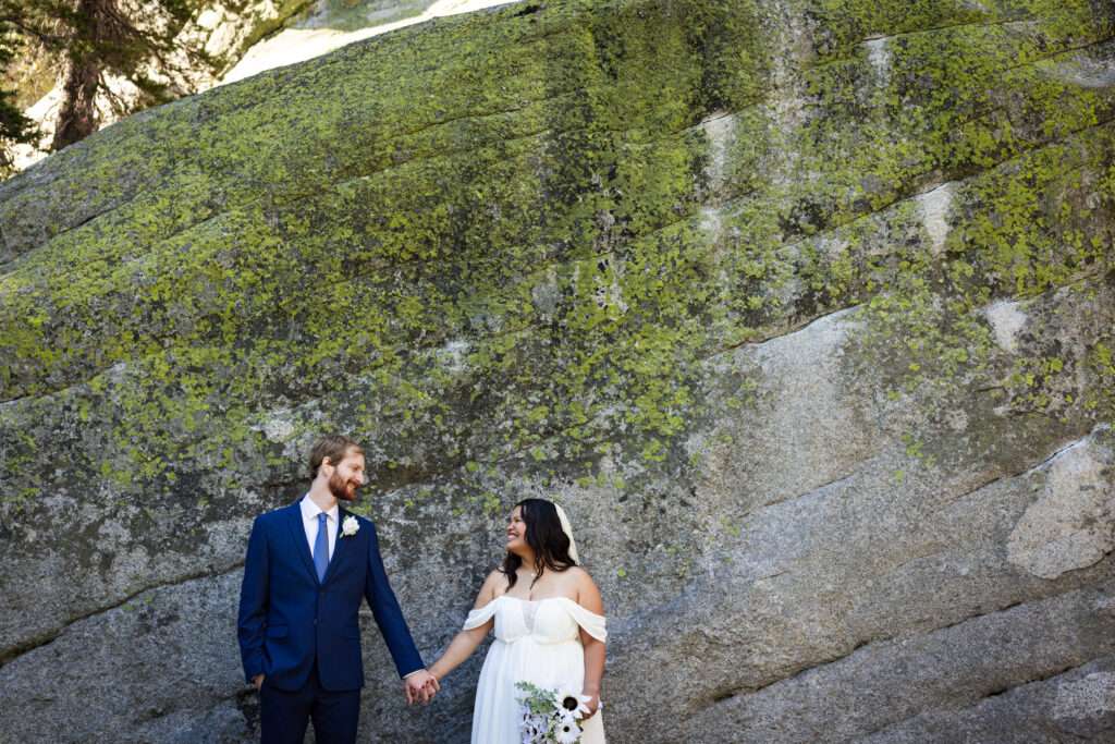 Newlyweds holding hands beside a moss-covered granite wall