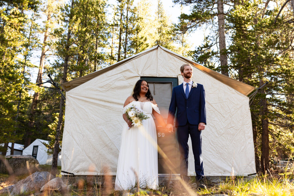 Newlyweds standing outside a canvas tent at a forest campsite