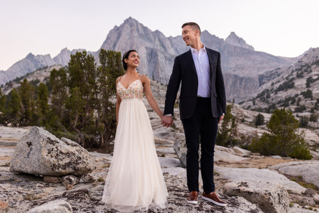 Newlyweds holding hands on a granite plateau beneath mountain peaks