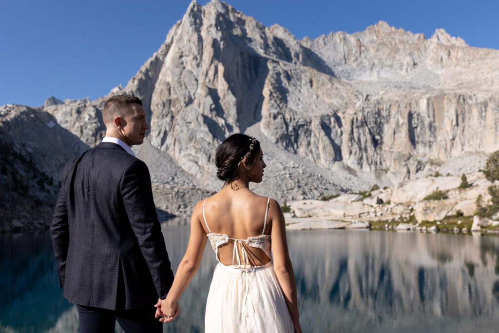 Newlyweds holding hands and looking across the lake toward granite peaks