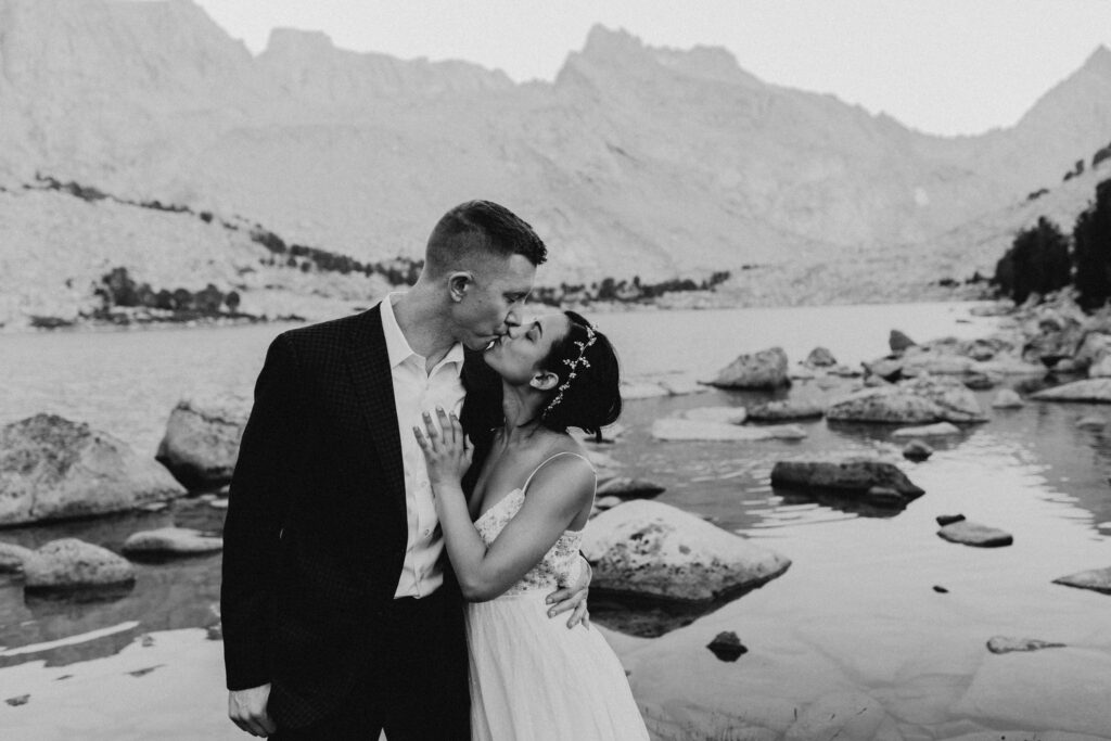 Black-and-white portrait of newlyweds kissing beside a mountain lake